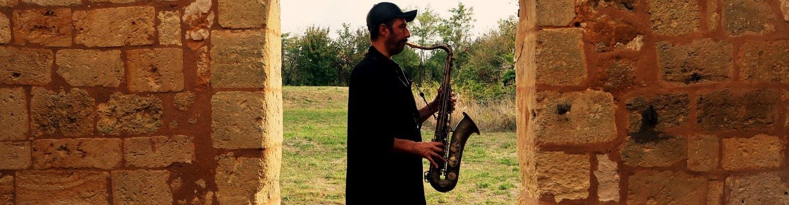 Ash Belgeri jouant du ténor dans l'embrasure d'une ruine de pierre, la campagne derrière