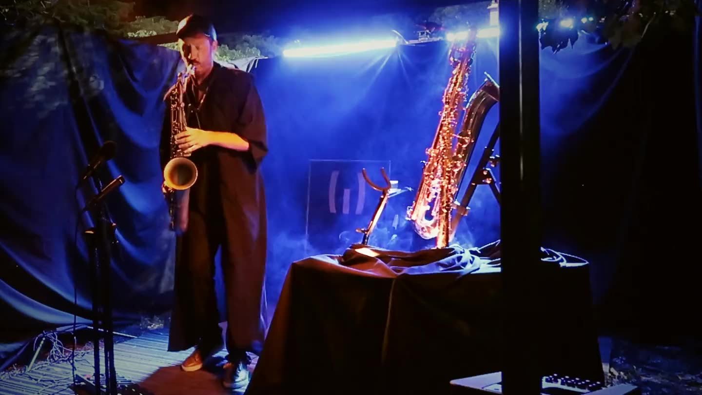Ash Belgeri joue du saxophone ténor sur scène, la nuit, les trois saxophones éclairés derrière lui